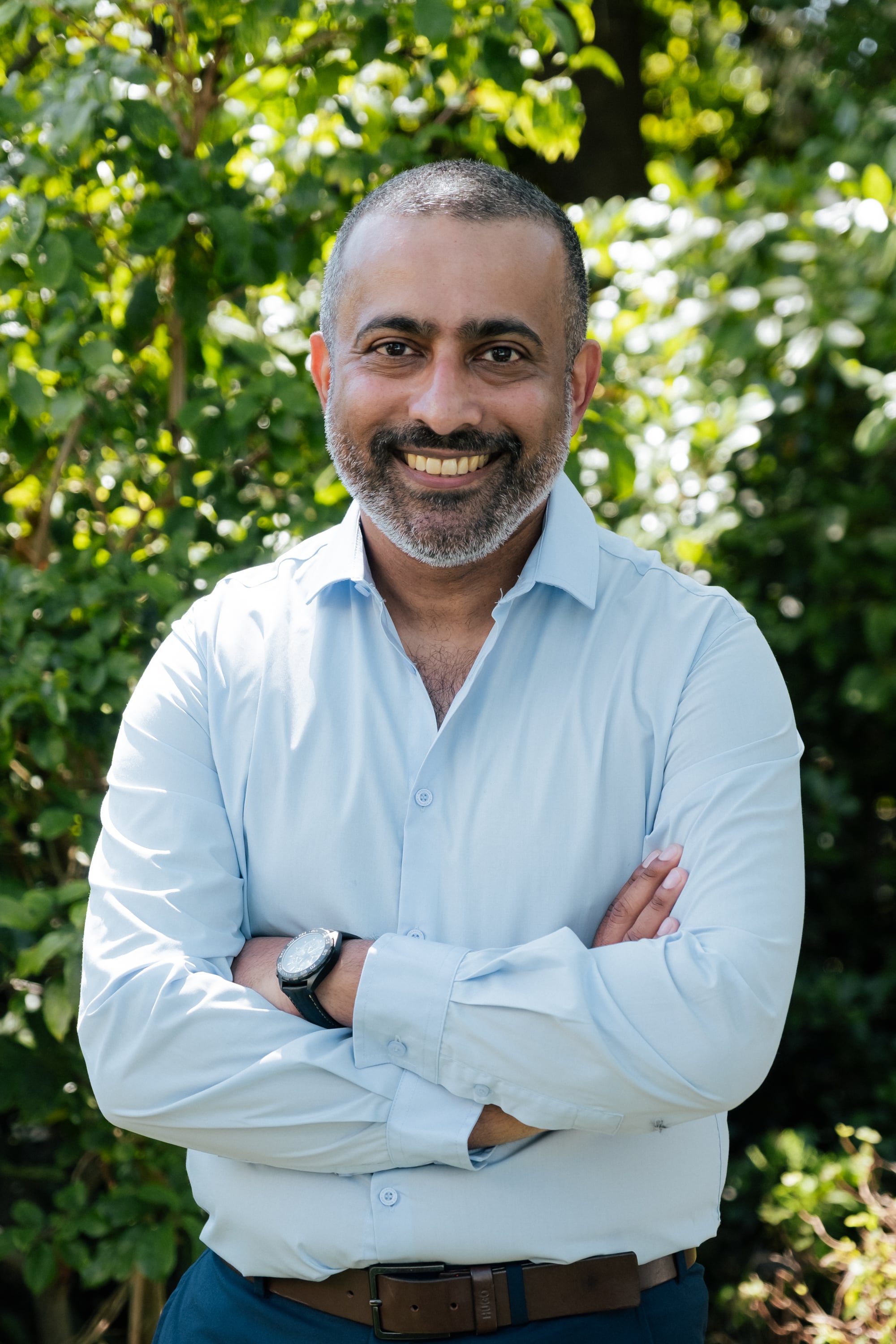 Dr Anith Chacko outdoors in a light blue shirt, arms crossed
