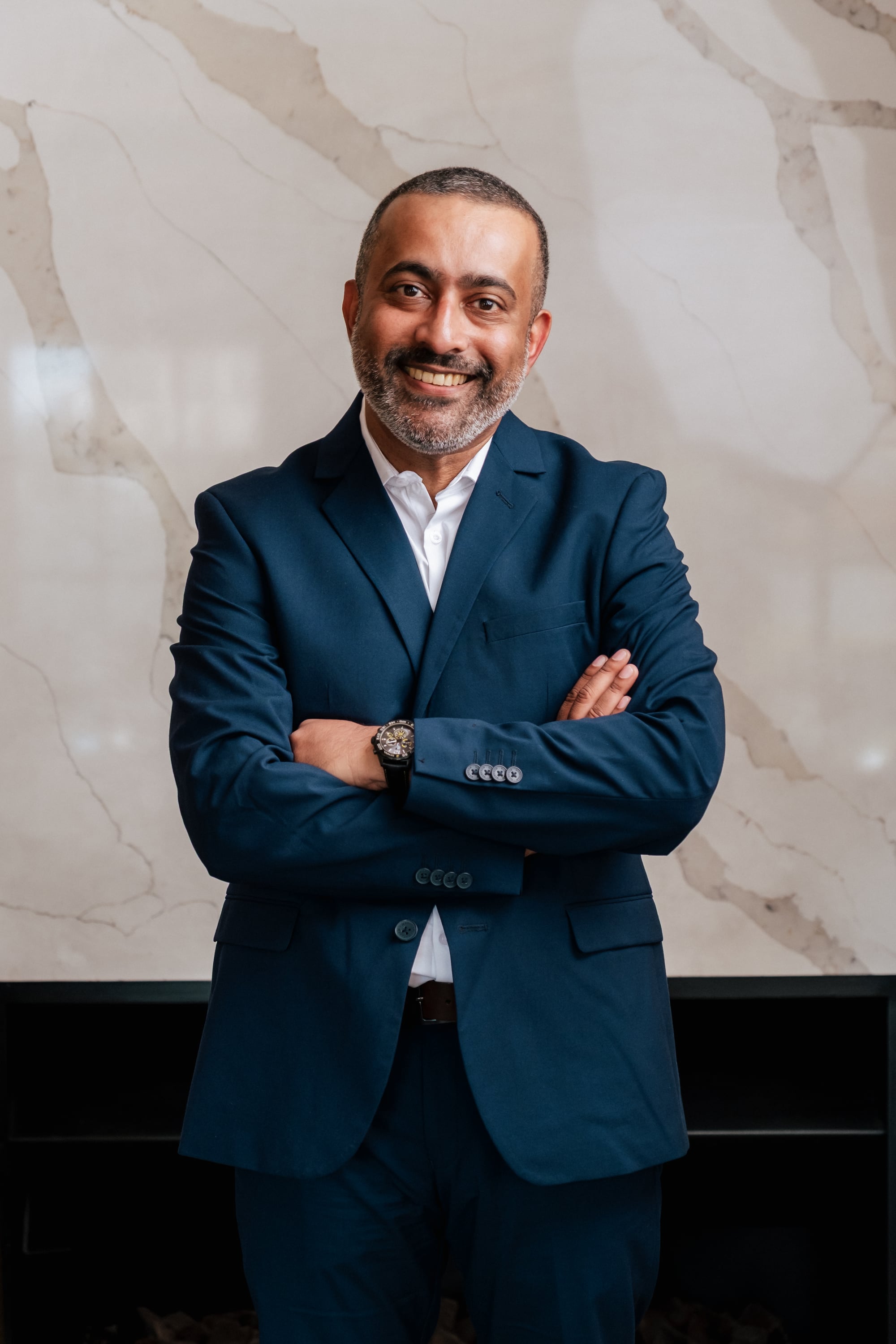 Dr Anith Chacko in a navy suit, arms crossed, against a marble wall